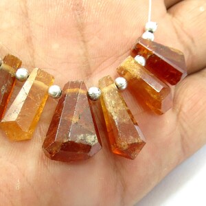May include: A strand of faceted, amber-colored gemstones with silver-colored beads. The gemstones are various shapes, including rectangular and trapezoidal, and are strung on a thin, white cord. The gemstones are held in a hand.