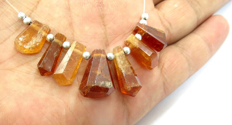 May include: A strand of faceted, amber-colored gemstones with silver-colored beads. The gemstones are various shapes, including rectangular and trapezoidal, and are strung on a thin, white cord. The gemstones are held in a hand.