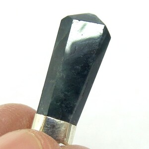 May include: A polished, black gemstone with a silver-colored metal band at the base. The gemstone has a rectangular shape with a faceted, angled top. The stone is held between fingers against a white background.