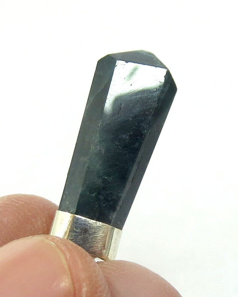May include: A polished, black gemstone with a silver-colored metal band at the base. The gemstone has a rectangular shape with a faceted, angled top. The stone is held between fingers against a white background.