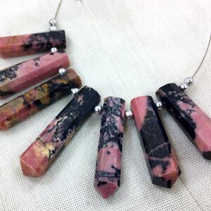 May include: A necklace featuring faceted, elongated gemstone beads. The beads are primarily pink with black and brown veining, strung on a thin silver wire. Each bead is approximately 1 inch long. The necklace is displayed on a white fabric surface.