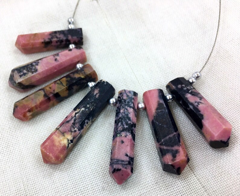 May include: A necklace featuring faceted, elongated gemstone beads. The beads are primarily pink with black and brown veining, strung on a thin silver wire. Each bead is approximately 1 inch long. The necklace is displayed on a white fabric surface.