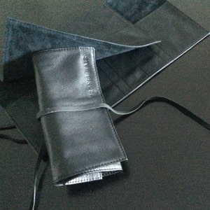 Puede incluir: Un estuche enrollable de cuero negro con el texto "ROBERTA PIERI" grabado en relieve en la parte delantera. El estuche se cierra con una correa de cuero negro y se encuentra sobre una superficie gris oscuro.