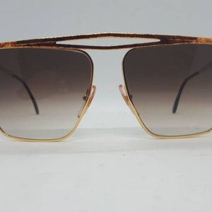 Pu&ograve; includere: Occhiali da sole dorati con montatura quadrata e lenti marroni sfumate.