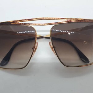 Pu&ograve; includere: Occhiali da sole dorati con lenti marroni e un design della montatura geometrico unico.