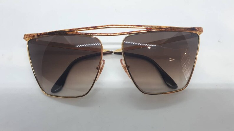 Pu&ograve; includere: Occhiali da sole dorati con lenti marroni e un design della montatura geometrico unico.