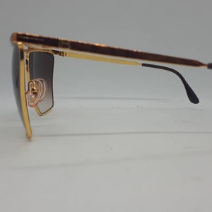 Pu&ograve; includere: Occhiali da sole dorati con lenti marroni sfumate e aste nere. Gli occhiali hanno una montatura quadrata.