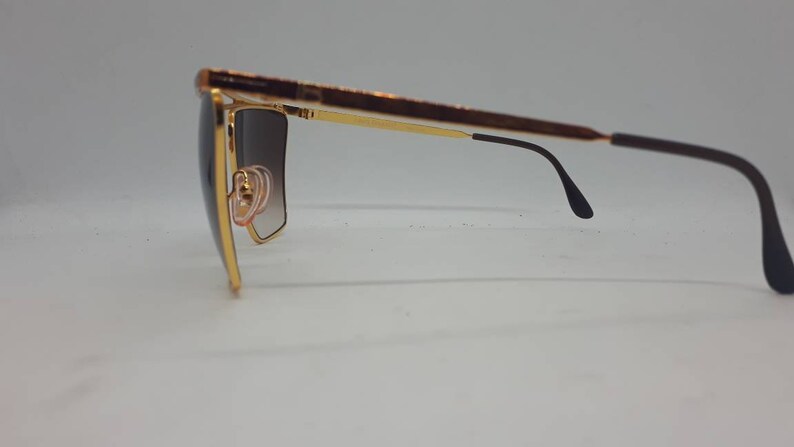 Pu&ograve; includere: Occhiali da sole dorati con lenti marroni sfumate e aste nere. Gli occhiali hanno una montatura quadrata.