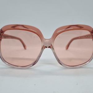 Könnte beinhalten: Übergroße, vintage Sonnenbrille in hellem Rosa. Die Rahmen sind ein durchscheinendes, warmes Rosa, und die Gläser haben einen passenden Farbton. Die Brille hat eine abgerundete, quadratische Form, und die Bügel sind dünn und gebogen.