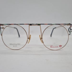 Könnte beinhalten: Vergoldete 24-Karat-Brille mit floralem Muster auf dem oberen Rahmen und einem Notendetail auf der rechten Seite. Die Brille hat runde Gläser und der Markenname "Casanova" ist auf dem rechten Glas aufgedruckt.