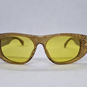 Könnte beinhalten: Goldfarbene Sonnenbrille mit gelben Gläsern. Die Sonnenbrille hat ein strukturiertes, fast schlangenhautähnliches Muster auf den Bügeln.