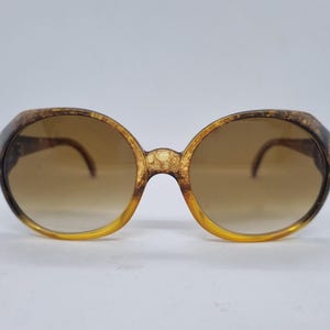 May include: Oversized round sunglasses featuring a mottled brown and gold tortoiseshell pattern on the frames. The lenses have a gradient tint, transitioning from a darker brown at the top to a lighter, translucent amber hue at the bottom.