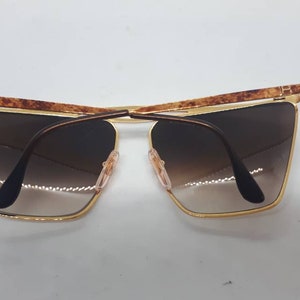 Pu&ograve; includere: Occhiali da sole dorati con aste in tartaruga marrone e lenti marroni sfumate.