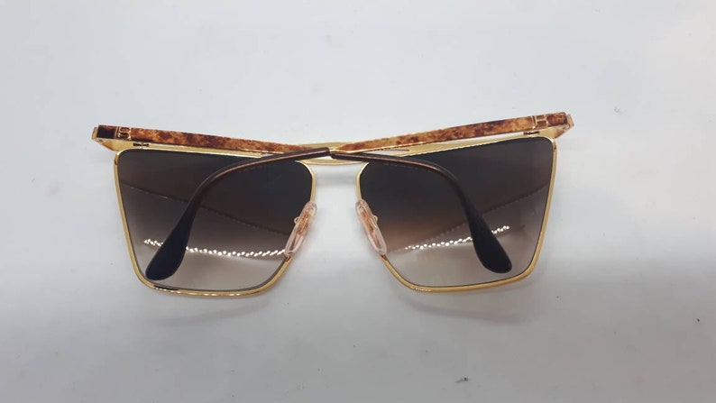 Pu&ograve; includere: Occhiali da sole dorati con aste in tartaruga marrone e lenti marroni sfumate.