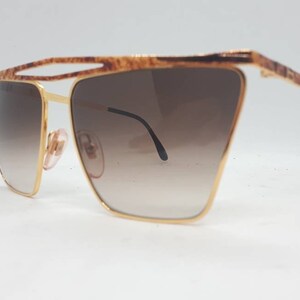 Pu&ograve; includere: Occhiali da sole dorati con lenti marroni sfumate e montatura tartaruga.