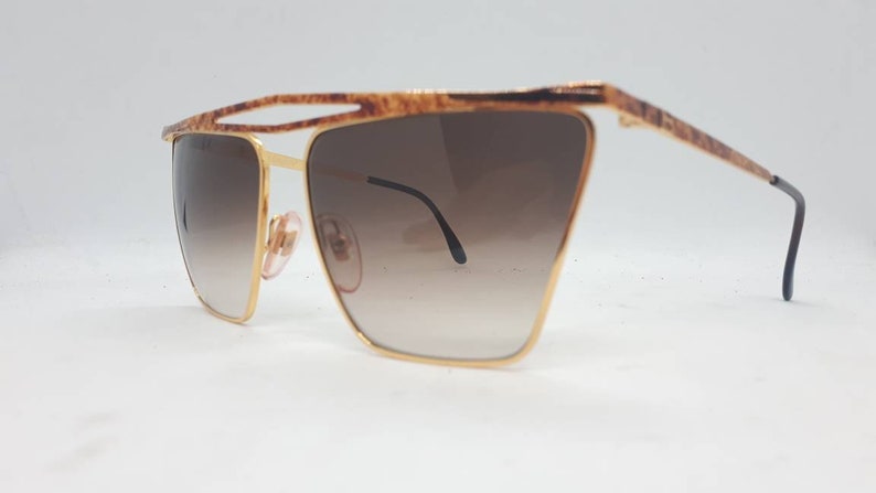 Pu&ograve; includere: Occhiali da sole dorati con lenti marroni sfumate e montatura tartaruga.