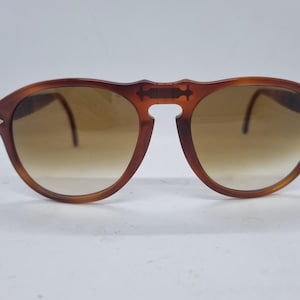 May include: Brown aviator sunglasses with gradient lenses. The sunglasses have a distinctive bridge design and silver accents on the temples. The lenses transition from a darker shade at the top to a lighter shade at the bottom.