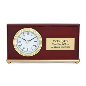 May include: A mahogany desk clock with a gold trim and a white face. The clock has a gold plaque with the text "Vicki Sykes, Chief Fun Officer, Allendale Day Care".