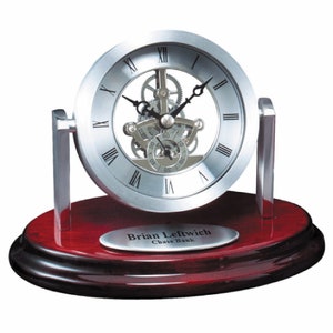 May include: Silver desk clock with a mahogany base. The clock face has Roman numerals and a skeletonized movement. The base has a silver plaque with the text "Brian Leftwich Chase Bank."