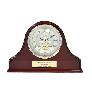 May include: A mahogany wood desk clock with a gold-toned face and Roman numeral hour markers. The clock has a visible skeleton movement and a personalized plaque that reads "Florence Bolak, Head Nurse, McGuff Nursing Home".