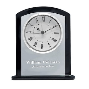 May include: A black and silver desk clock with a round clock face featuring Roman numerals. The clock has the text "William Coleman Attorney at law" engraved on the front.