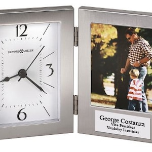May include: A silver-toned picture frame with a clock on the left side and a photo on the right. The clock face is white with black hands and numbers. The photo shows a man and a boy in a park. The frame has a nameplate that reads "George Costanza, Vice President, Vandelay Industries."