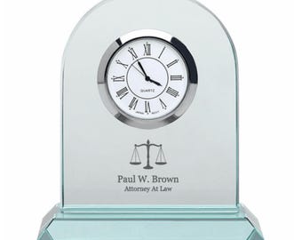 Può includere: Un orologio da tavolo in vetro con un quadrante argentato e indici delle ore con numeri romani. L'orologio ha un simbolo di bilancia della giustizia e il testo "Paul W. Brown Attorney At Law" inciso sulla base.