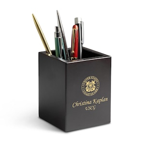 May include: A dark wooden pen holder with a gold Coast Guard emblem and personalized text. The holder contains several pens in various colours, including gold, green, red, and silver. A professional desk accessory.