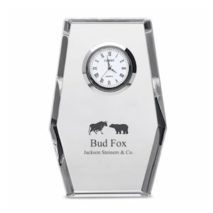 May include: A clear glass desk clock with a silver face and black hands. The clock features a bull and bear silhouette with the text "Bud Fox Jackson Steinmen & Co."