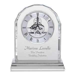 May include: A clear crystal clock with a silver face and black Roman numeral hour markers. The clock has a visible gear mechanism and is engraved with the text "Marissa Lavalle, Vice President, Vandelay Industries".