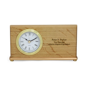 May include: A wooden desk clock with a gold trim around the face. The clock face has Roman numerals and a gold-colored hour and minute hand. The clock is engraved with the text "Peter V. Parker, Vice Principal, Albertus Magnus High School".