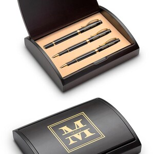 May include: A wooden pen set with three black pens with gold accents, presented in a dark wooden box. The pens are engraved with the name "James Miller". The box lid features a gold logo with the name "Miller".