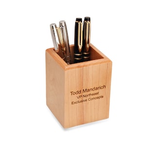 May include: A square wooden desk tidy with a light brown finish. The tidy has an engraved name and title: "Todd Mandarich, VP Northeast, Exclusive Concepts". Three pens are in the tidy.