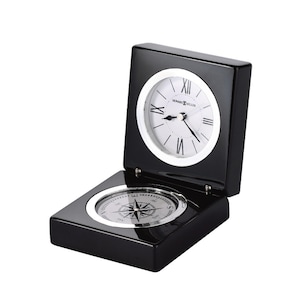 May include: A black wooden clock with a compass on the bottom. The clock face is white with black Roman numeral hour markers and black hands. The compass is silver with a black needle. The clock is made by Howard Miller.