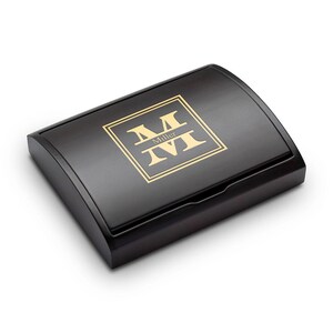 Pu&ograve; includere: Una scatola di legno nera con coperchio curvo. La scatola presenta un monogramma dorato con il nome "Miller" al centro. Il monogramma &egrave; racchiuso in un quadrato dorato.
