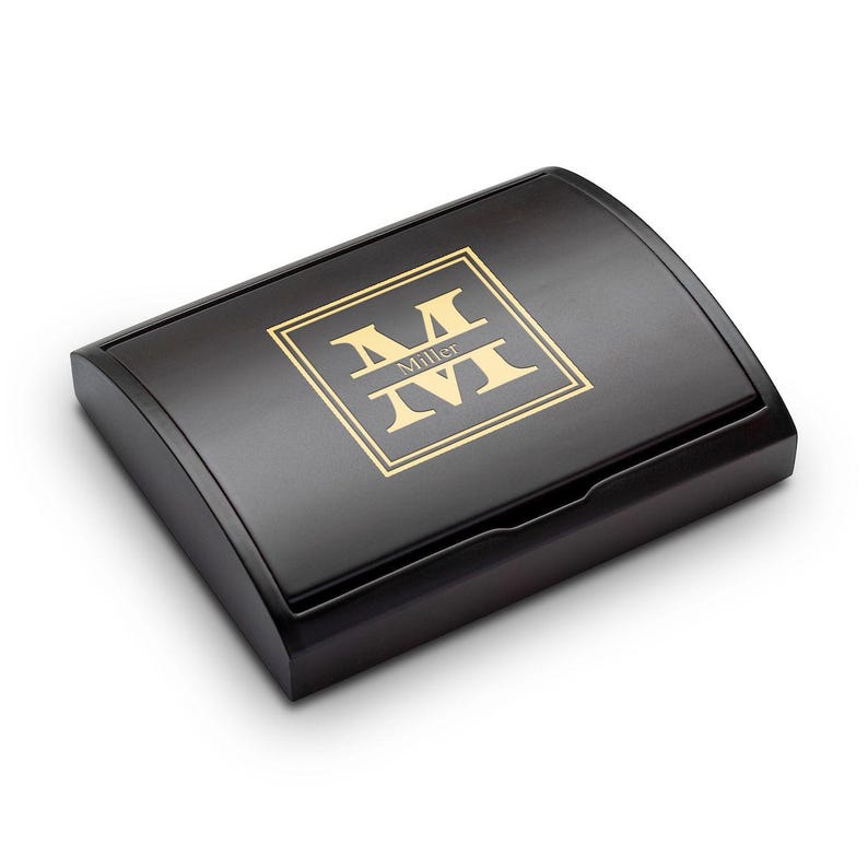 Pu&ograve; includere: Una scatola di legno nera con coperchio curvo. La scatola presenta un monogramma dorato con il nome "Miller" al centro. Il monogramma &egrave; racchiuso in un quadrato dorato.