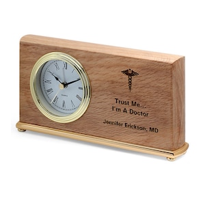 May include: A wooden desk clock with a gold trim and a white face. The clock has Roman numerals and a medical symbol engraved on the wood with the text "Trust Me... I'm A Doctor" and "Jennifer Erickson, MD".