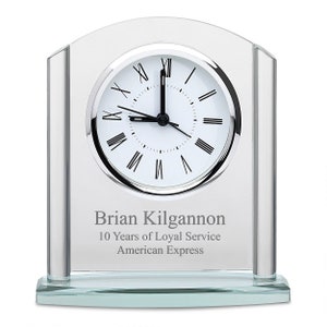 May include: A clear glass clock with Roman numeral markings on the face. The clock is on a glass base with a silver inscription that reads "Brian Kilgannon 10 Years of Loyal Service American Express".