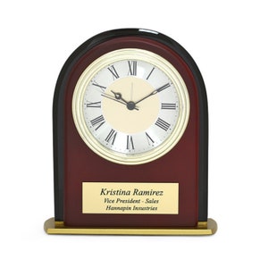 Peut inclure: Une horloge de bureau en bois d'acajou avec une bordure dorée et un cadran noir. L'horloge a des chiffres romains pour les heures et une base dorée. Une plaque personnalisée indique "Kristina Ramirez, Vice-présidente - Ventes, Hannapin Industries".