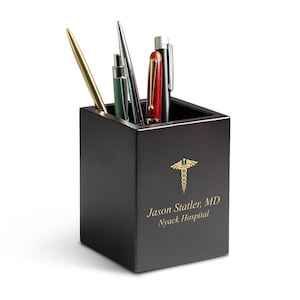 May include: A black wooden desk organiser with a gold medical symbol and the text "Jason Statler, MD Nyack Hospital". The organiser has four pens inside.