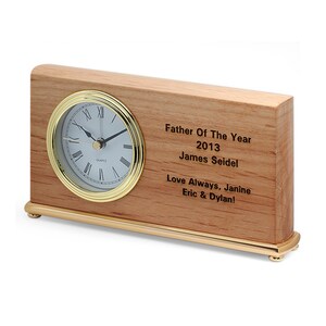 May include: A wooden desk clock with a gold trim and a white face. The clock has a personalized inscription that reads "Father Of The Year 2013 James Seidel Love Always, Janine Eric & Dylan!"