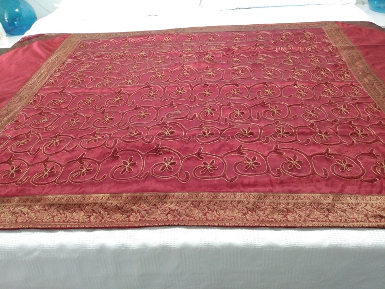 以下が含まれることがあります： 金色の花の刺繍が施された、バーガンディ色の織物。生地は、様式化された花とつるの全面的なパターンを特徴としています。繰り返しのデザインの装飾的なボーダーが端を縁取っています。この織物は、装飾用のスローまたはテーブルクロスである可能性があります。