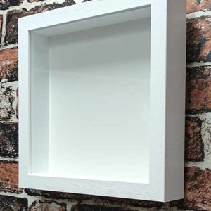 May include: A white shadow box frame, rectangular in shape, with a recessed interior. It's mounted on a brick wall with brown, red, and grey bricks. The frame's interior is also white.
