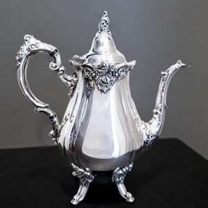 May include: A silver-colored teapot with ornate detailing. The teapot features a curved handle, a long spout, and a decorative lid. The base has three small feet. The design includes floral and scrollwork patterns, giving it a vintage aesthetic.