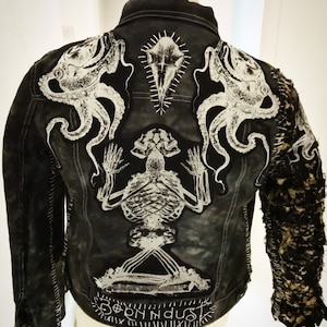 May include: Black denim jacket with white embroidered octopus, skeleton, and skull designs. The jacket has a distressed look with frayed edges and stitching. The text "Born in Dust" is embroidered on the bottom.