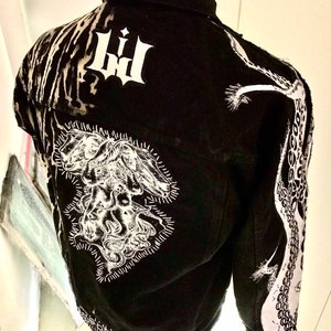 以下が含まれることがあります： 白いグラフィックデザインが施された黒いデニムジャケット。ジャケットの背面には、触手のある生き物の大きな白いグラフィックが、左袖には触手のある生き物の小さな白いグラフィックが描かれています。ジャケットには、背面に「LH」の白いグラフィックも描かれています。