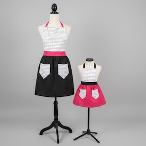 以下が含まれることがあります： マネキンに2つのエプロン。大きなエプロンは、スクリプト付きの白いトップス、ピンクのネックストラップとウエストバンド、白いポケットのディテールが付いた黒いスカートです。小さいエプロンはピンクで、黒いウエストバンドと白いポケットのディテールが付いています。