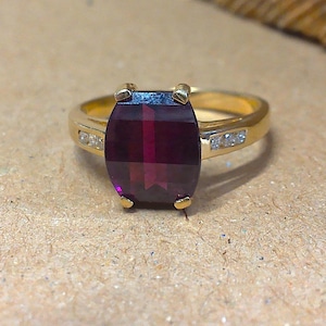 May include: A gold ring featuring a large, rectangular, deep red gemstone. The ring has small, clear accent stones on each side of the main stone. The band is a simple, polished gold.