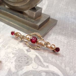 Peut inclure: Une broche en or avec un design délicat orné de pierres précieuses rouges et d'accents de perles.