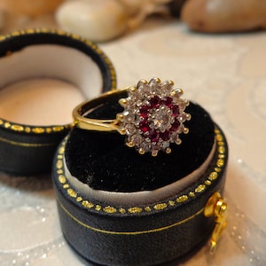 以下が含まれることがあります： 黒いリングボックスに入った、大きな赤と白の宝石が付いた金のリング